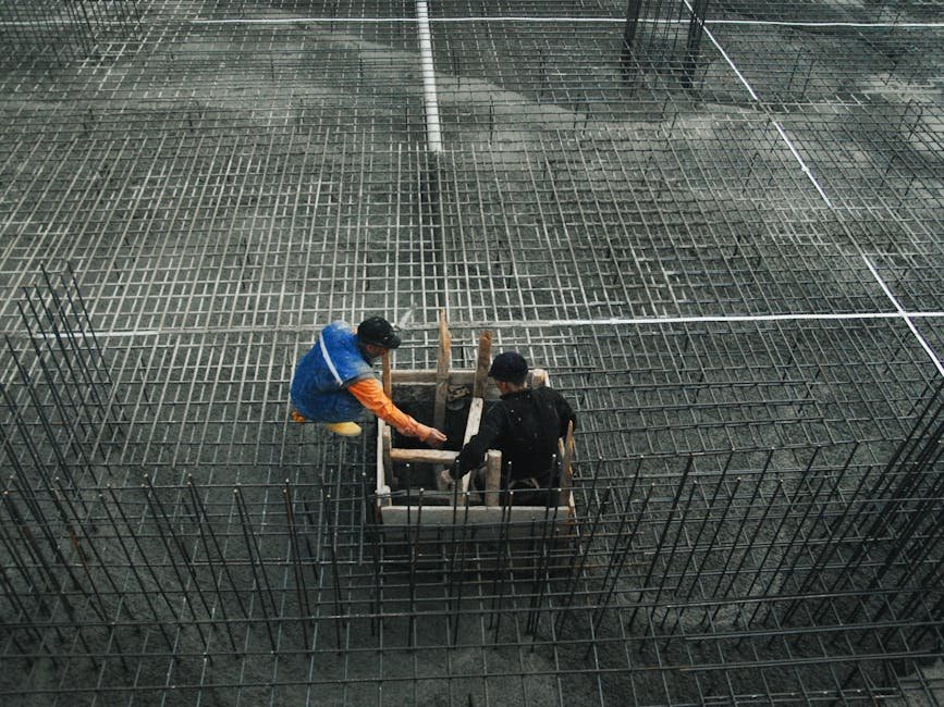 Two workers operating on a construction site with steel frameworks and concrete. High-angle view captures teamwork and industry.